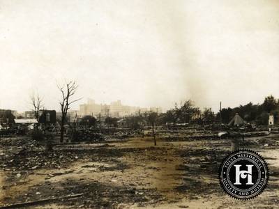 Fields of Despair - Some spaces were so decimated nothing but empty fields were left after the violence subsided. In this Greenwood District photo of the ruins, a tent is visible at the far right. On the left several African Americans stand underneath a shed with a metal roof.
