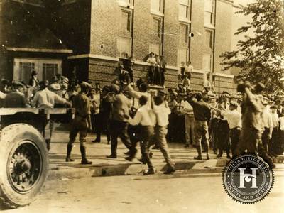 A Plague of Violence - It is unclear how many Black people were arrested, but whites who were deputized by law enforcement, along with the Oklahoma National Guard jailed many of them, blaming them for the violence. Here a group of African American men being led into Convention Hall holding their hands in the air. Many of the white men in the photo are holding guns, and some are watching from the windowsills of the building.