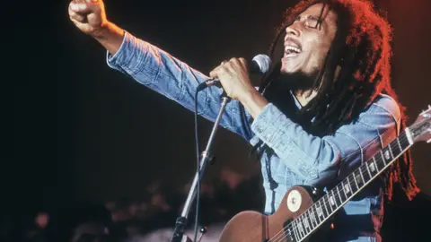 Bob Marley performs on stage, in a concert at Grona Lund, Stockholm, Sweden. He extends his fist as he sings into the microphone, with an electric guitar. (Photo by Hulton Archive/Getty Images)