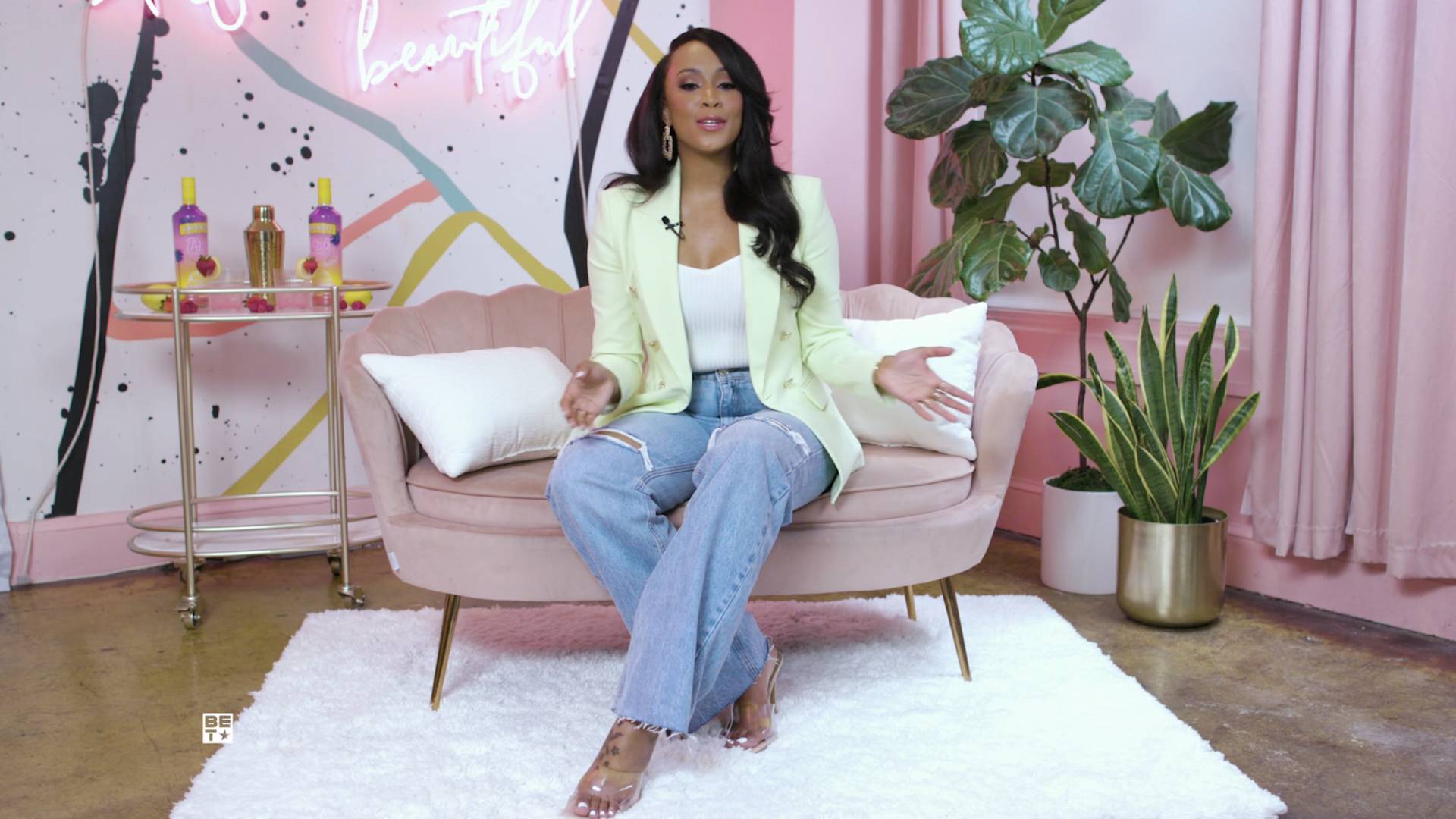 Amanda Booz in jeans, a white shirt and neon yellow blazer, sitting on a pink chair with a colorful background.