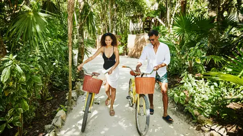 Couple walking bikes on pathway at tropical resort.