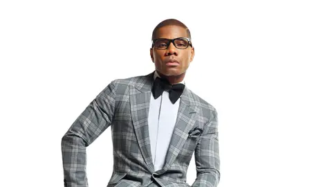 Kirk Franklin posing in a grey suit, white shirt, and black bowtie against a white background.