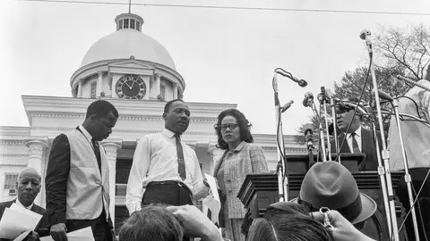View of American religious and Civil Rights leaders John Lewis (in vest) and Martin Luther King Jr (1929 - 1968) and his wife, Coretta Scott King (1927 - 2006), on the podium before the Selma to Montogomery March rally on the steps on the Alabama State Capitol, Montgomery, Alabama, March 25, 1965. Also visible is union leader A Philip Randolph (1889 - 1979) (seated at left). The Confederate and Alabama flags fly over the Capitol. (Photo by Charles Shaw/Getty Images)