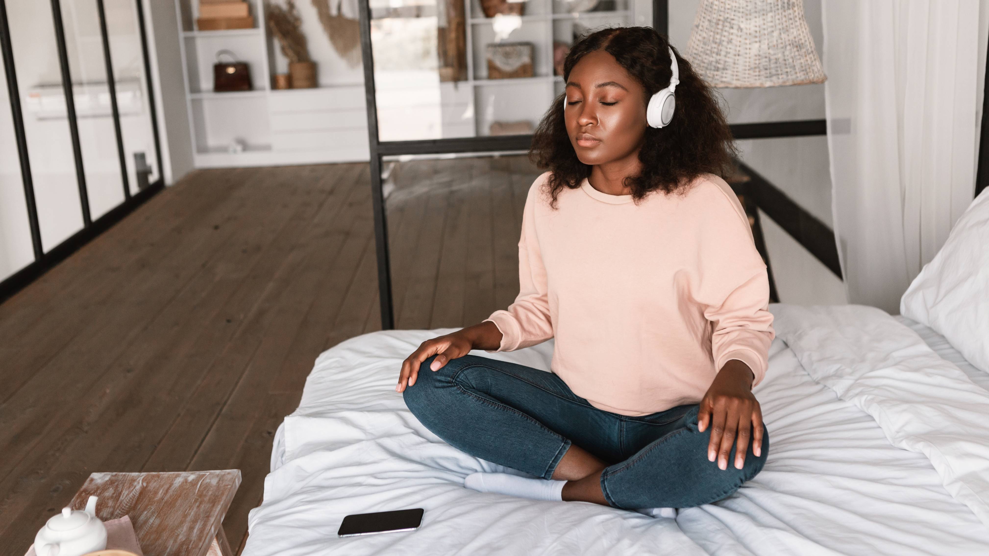 Relaxed African American Lady Meditating Wearing Headphones In Bedroom.