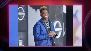 Hip Hop artist Bleu smiling on a red carpet wearing a blue jacket, black shirt, and several chain necklaces.