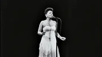 Legendary artist Anita Baker in an older black and white photo, performing on stage in a white dress.