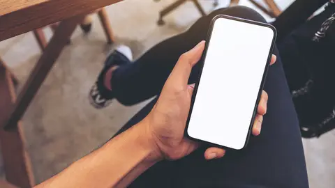 Top view mockup image of a man's hand holding white mobile phone with blank desktop screen on thigh while sitting in cafe