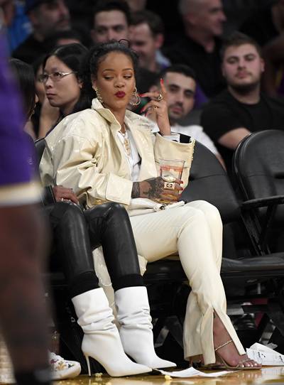 RiRi With A Serving Of Side Eye - We love a courtside RiRi and we know she was spilling some serious tea to her bff, Melissa Forde,with this look! Her nude, courtside look featured a SS19 couture Alexandre Vauthier, leather, trench coat; FENTY, leather, skinny pants with ankle slits ($1,340); Amina Muaddi 'Gilda' ankle strap sandals ($945); and a mini croc, white, Fendi bag. (Photo: Kevork S. Djansezian/Getty Images)