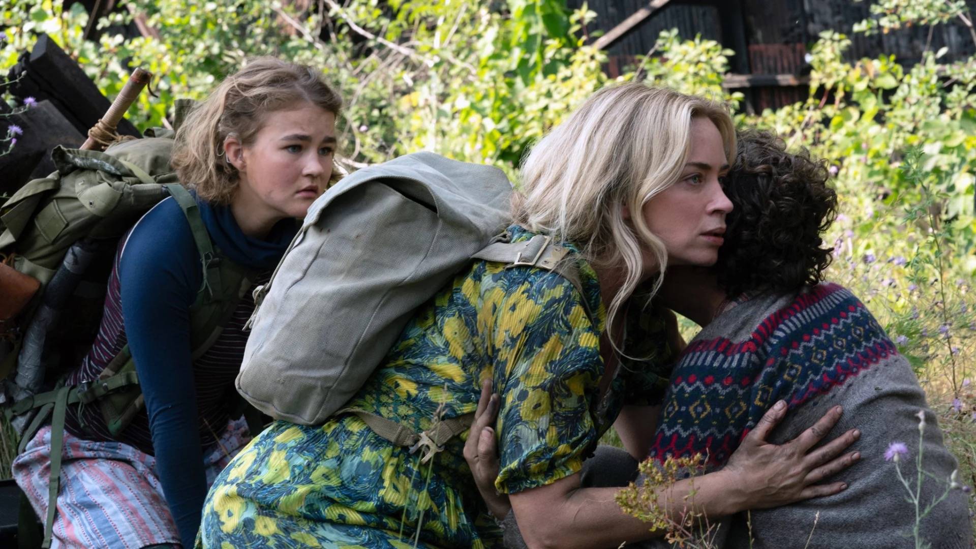 Emily Blunt crouching in fear in floral address and backpack holding a small boy in a grey jumper. A young girl wearing a backpack and navy top is crouching behind her 