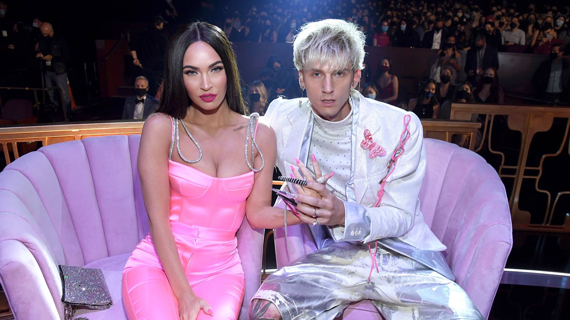 Megan Fox in pink jumpsuit sitting next to Machine Gun Kelly in silver short suit. The two have their hands entangled and are sitting on a plush light purple sofa 