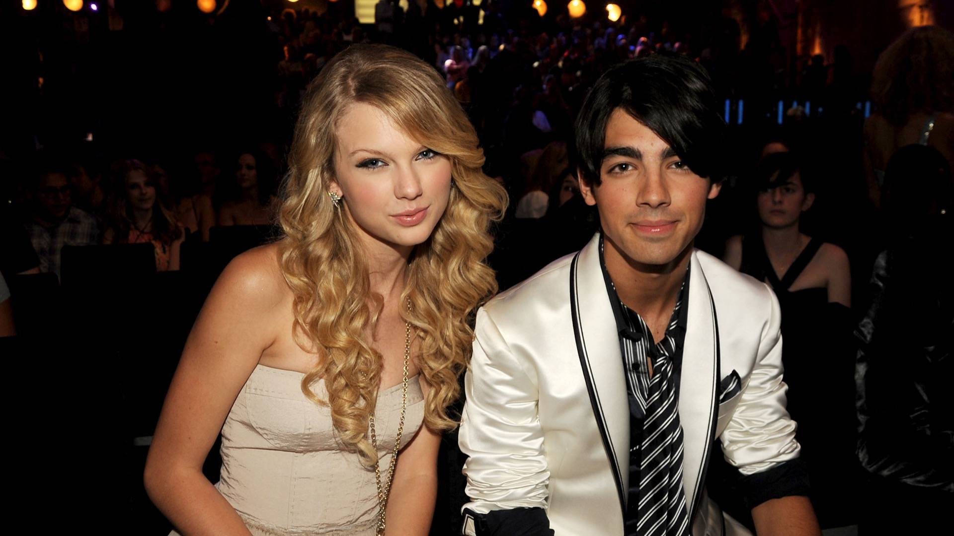 Taylor Swift in beige dress sitting next two Joe Jonas in silk cream jacket and striped tie 