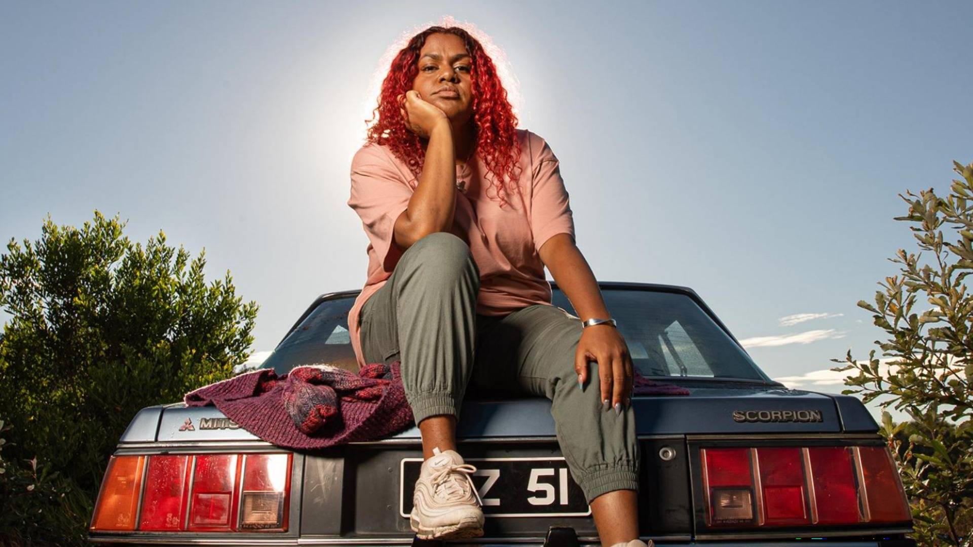 Miiesha with red hair sitting on boot of old blue Mitsubishi with chin resting on hand 