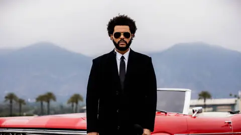 The Weeknd poses in front of red sports care in black suit, dark tie and aviator sunglasses