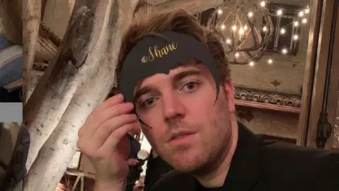 Shane Dawson holding cardboard bat-shaped placeholder with the word 'Shane' on it on forehead