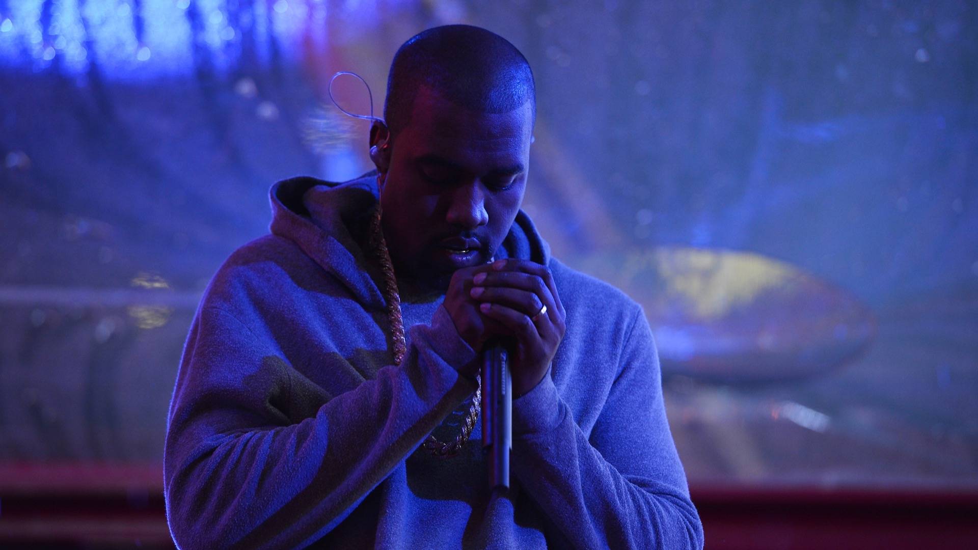 Kanye West holding microphone in grey hoodie and chain necklace, with dim lighting and multicoloured background 