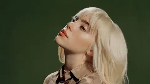 Billie Eilish looking off camera with blonde hair and beige shirt in front of dark green background