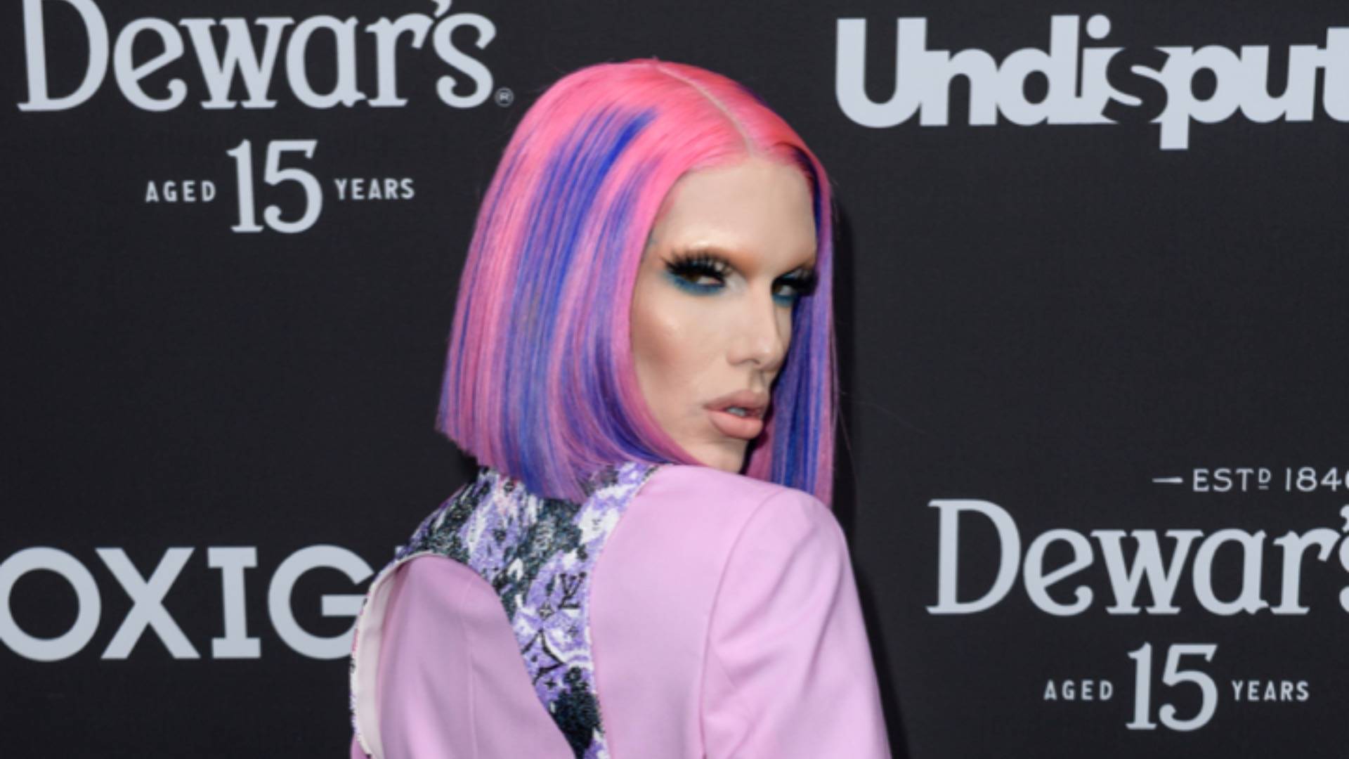 Jeffree Star posing in decorative back brace, lilac suit jacket and hair styled into a straight bob containing blue and pink streaks 