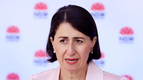 Gladys Berejeklian addresses media in front of NSW Health patterned wall in light pink blazer 