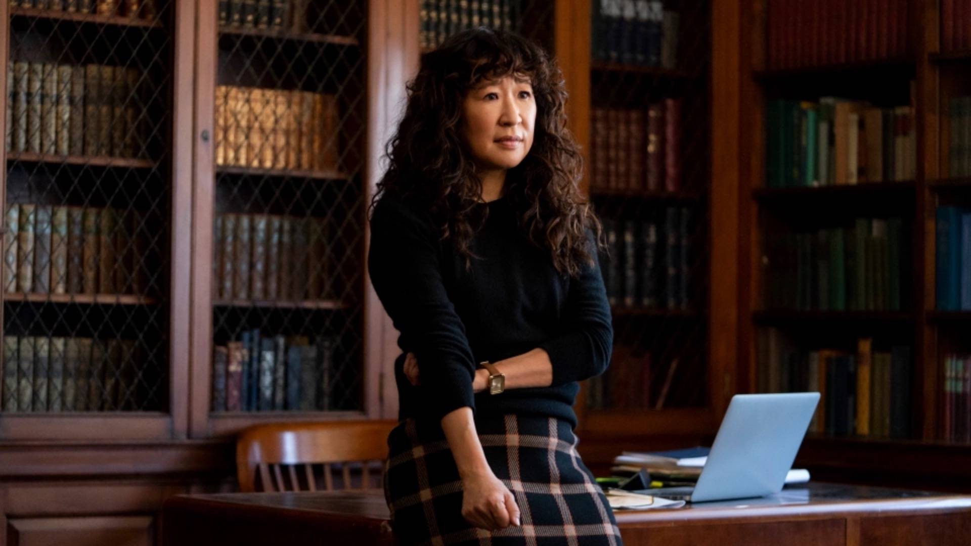 Sandra Oh in dark jumper and plaid skirt, leaning against desk in University office and surrounded by books in Netflix show 'The Chair'