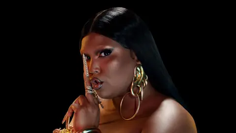 Lizzo posing with straight hair and gold nails and earrings, putting finger to mouth in 'shush' gesture
