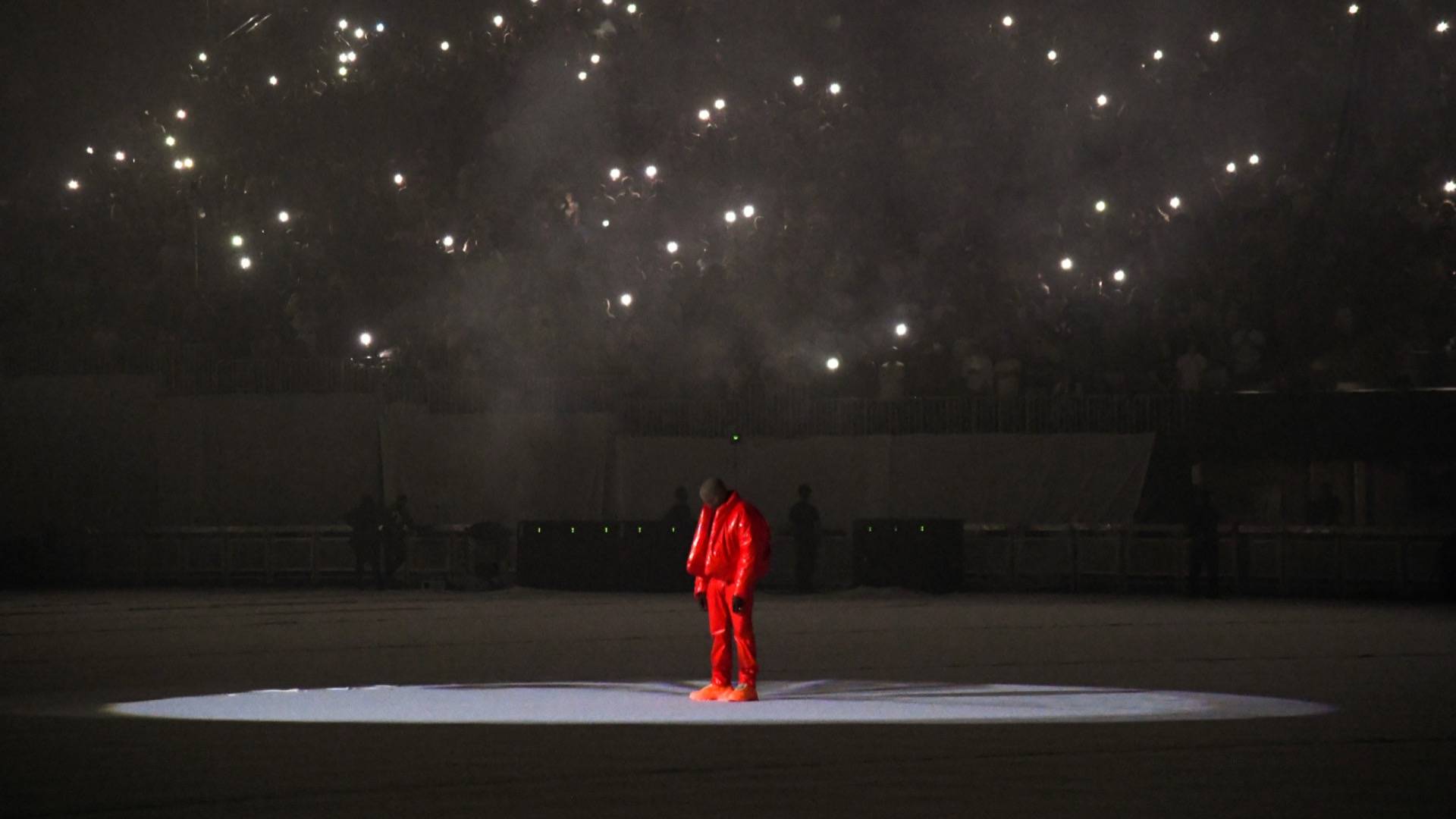 Kanye West in mask and red outfit on stage at DONDA listening party, with lights from the audience visible overhead