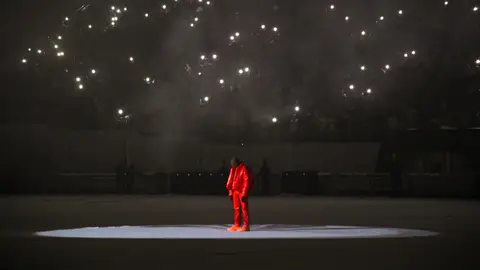 Kanye West in mask and red outfit on stage at DONDA listening party, with lights from the audience visible overhead