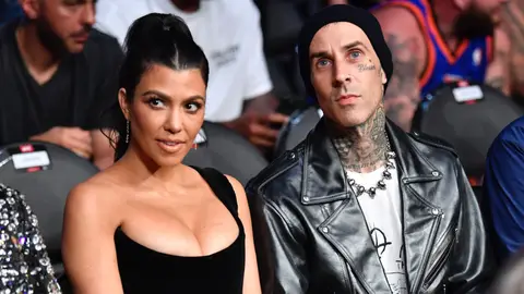 Kourtney Kardashian with hair in ponytail and dressed in a low-cut black top sitting next to Travis Barker in a white shirt, leather jacket and black beanie. 