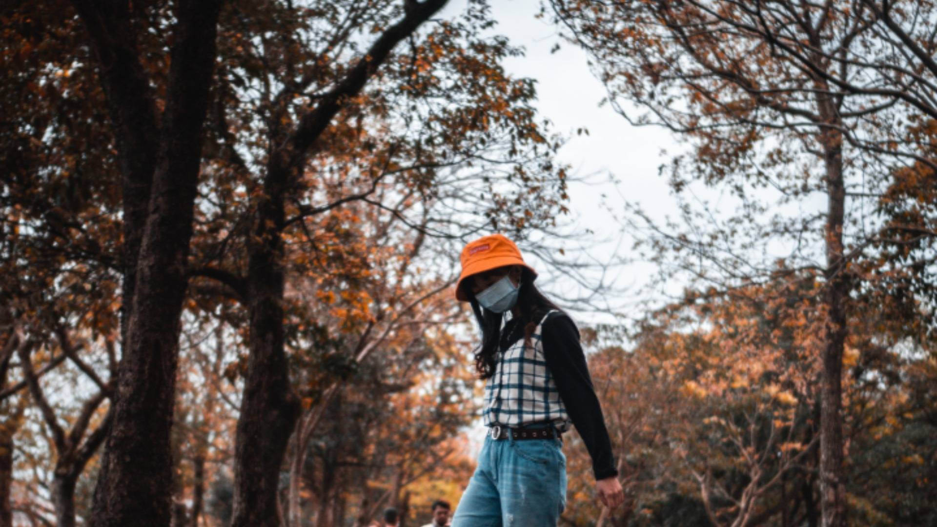 Girl in orange hat and face mask standing outdoors surrounded by autumnal foliage 