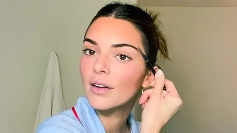 Kendall Jenner with hair up, wearing blue pyjamas and grooming eyebrows with spoolie in Vogue 'Beauty Secrets' video