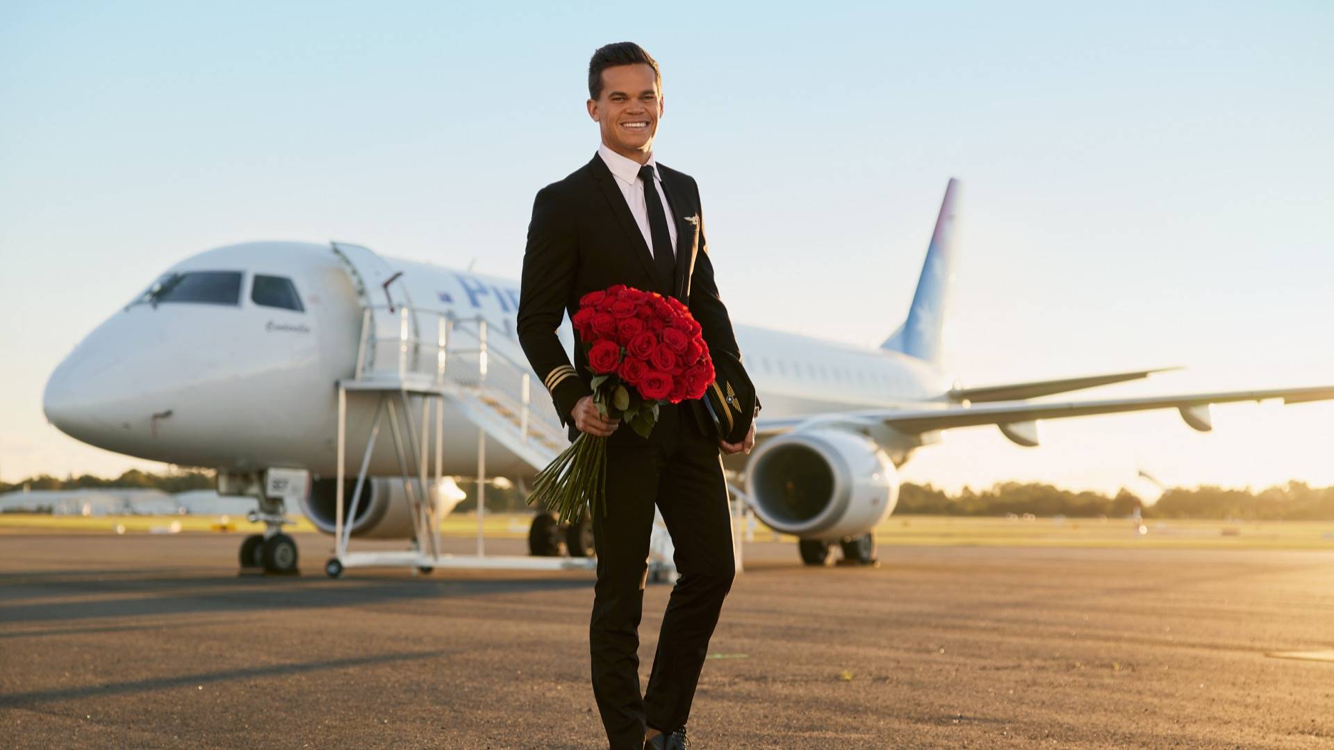 New Bachelor Jimmy Nicholson dressed in pilot's uniform and posing with bunch of red roses in front of aeroplane 