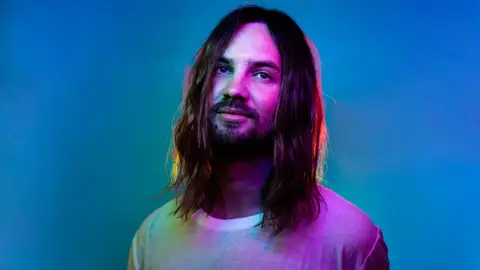 Tame Impala smiling with lights coloured in the background hair down with white tee