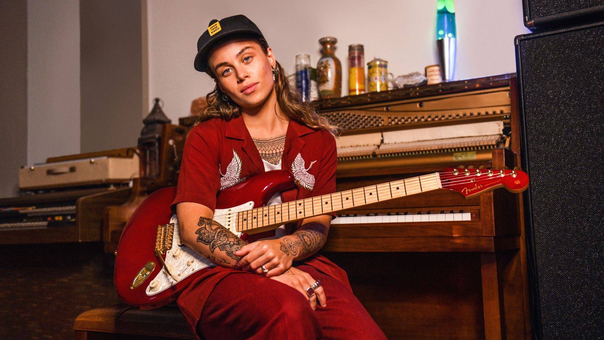 tash-sultana-in-red-suit-holds-fender-guitar-sits-over-piano-poses-for-camera