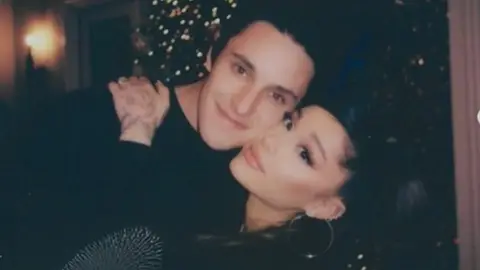 Ariana Grande with hands on Dalton Gomez's shoulders in front of Christmas tree