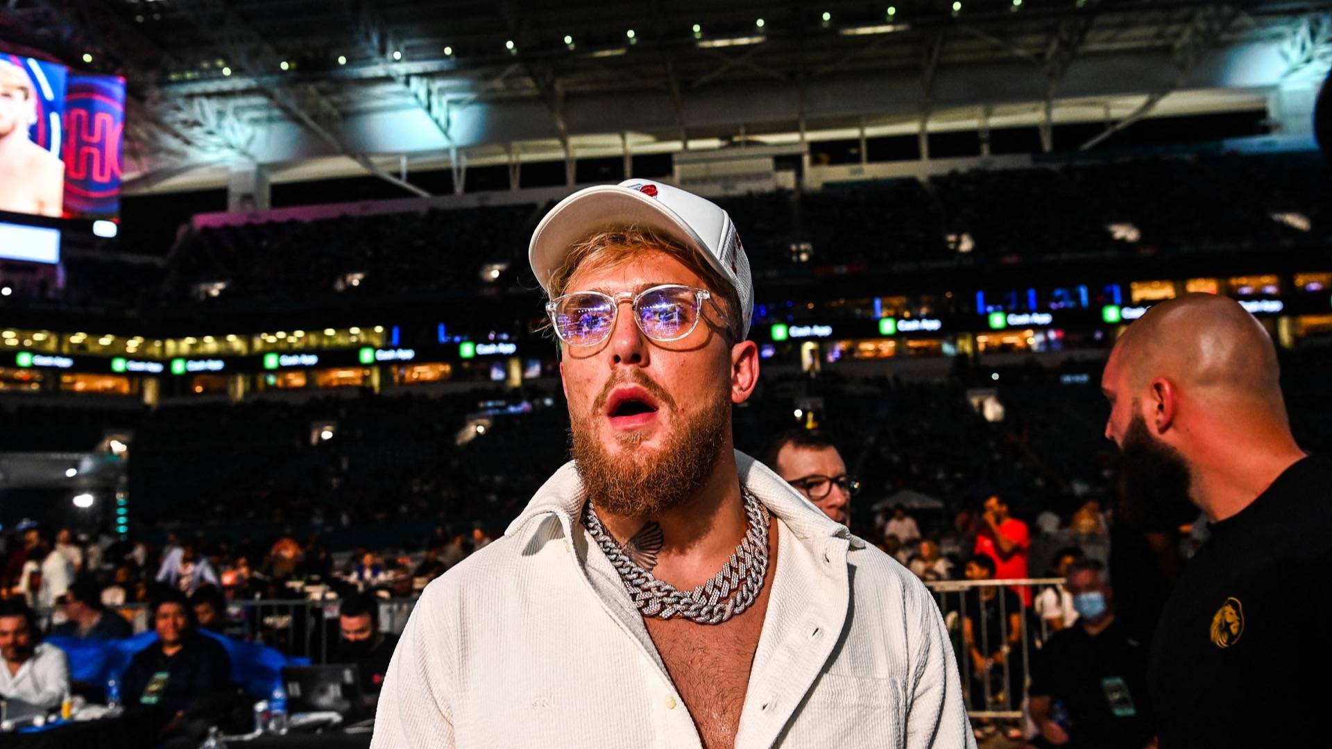 Jake Paul wearing glasses, white button down shirt and white baseball cap, looking off camera open-mouthed