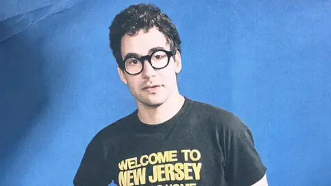 Jack Antonoff poses in front of blue screen in black and yellow shirt welcome to new jersey 