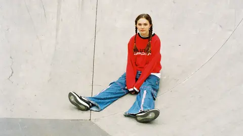 Sycco sitting in red jumper and blue jeans hair in plaits skate park