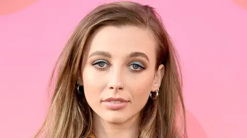 Emma Chamberlain posing with hair down and blue eyeshadow in front of pink background