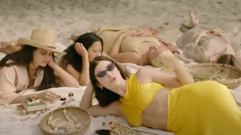Lorde lying on beach singing wearing sunglasses and long yellow skirt and yellow crop top with friends in the background, a still from her new 'Solar Power' video
