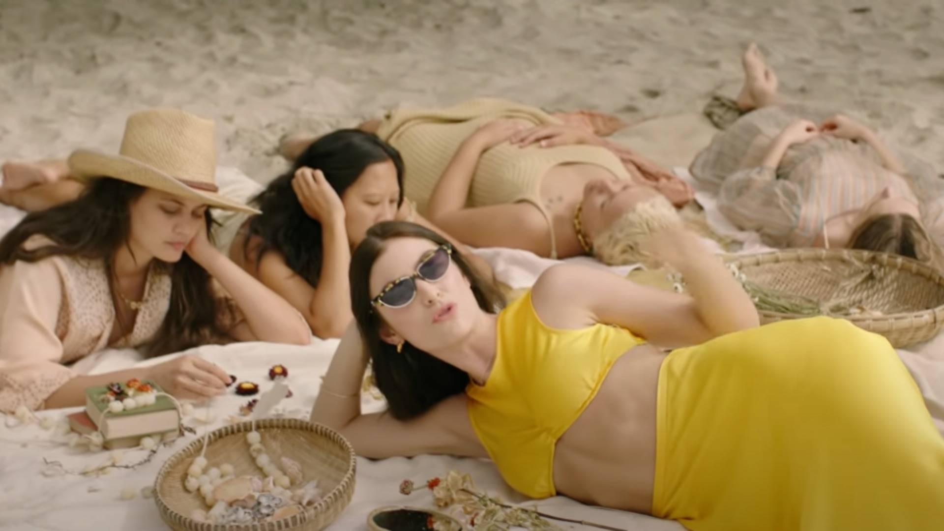 Lorde lying on beach singing wearing sunglasses and long yellow skirt and yellow crop top with friends in the background, a still from her new 'Solar Power' video