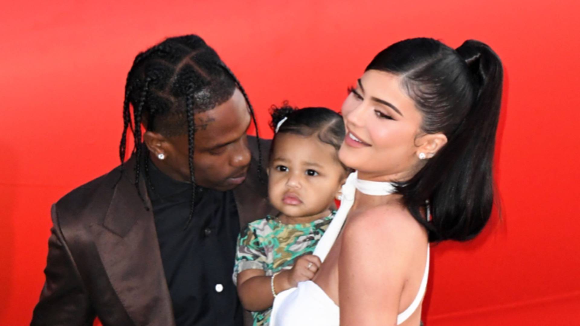 Travis Scott and Kylie Jenner posing with Stormi in front of red background