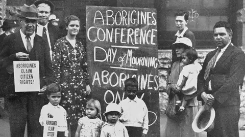 Black and white image of a group of people at First Day of Mourning in 1930s Australia. holding protest signs reading 'Aborigines claim citizen rights' and 'Aborigines conference, day of morning, Aborigines only.' From the left is William Ferguson, Jack Kinchela, Isaac Ingram, Doris Williams, Esther Ingram, Arthur Williams, Phillip Ingram, Louisa Agnes Ingram OAM holding daughter Olive Ingram, and Jack Patton. 