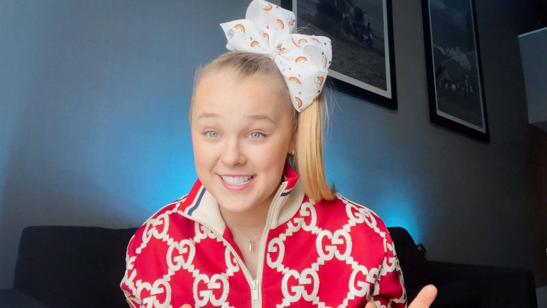 JoJo Siwa In red jumper zipped up and white rainbow bow on video call still smiling