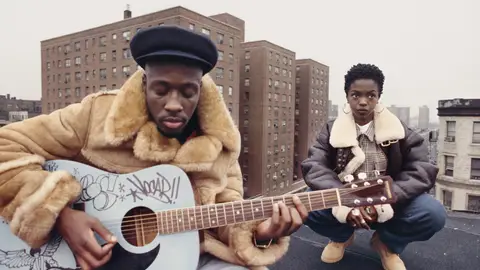 Wyclef Jean, wearing a brown fur coat and a cap, strums a baby blue guitar on a rooftop in East Harlem, New York. Behind him crouches Lauryn Hill.