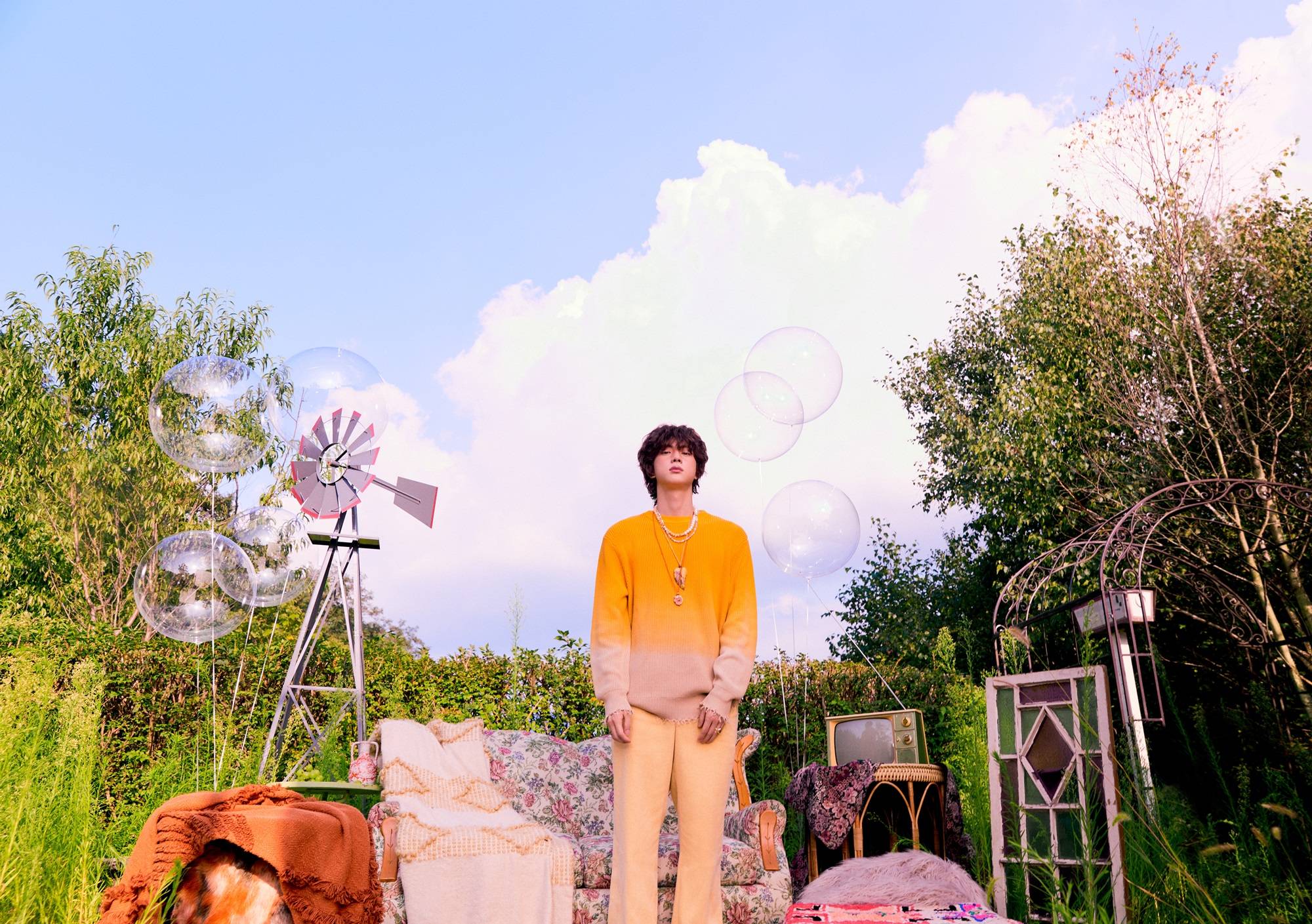 Jin of BTS poses in a sunny farm scene wearing a yellow shirt