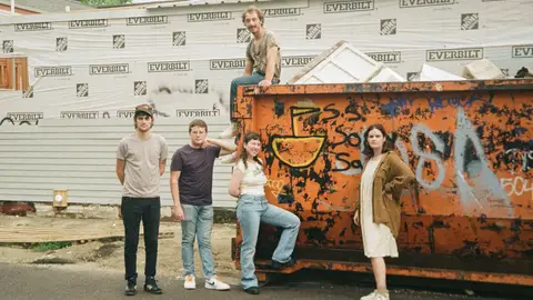 Wednesday pose in front of a graffiti'd dumpster