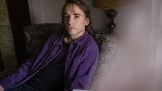 Andy Shauf poses in a purple shirt in an upholstered armchair