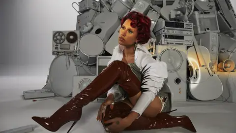 Raye thoughtfully poses in long leather boots in front of an artful mound of instruments and recording equipment