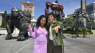 Anthony Ramos and Dominique Fishback pose in front of a squadron of Transformers characters