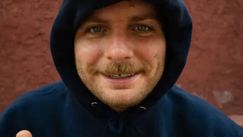 Mac DeMarco poses in a blue hoodie with a hat over his hood on his head reading "Mac's Record Label" while flashing a peace sign