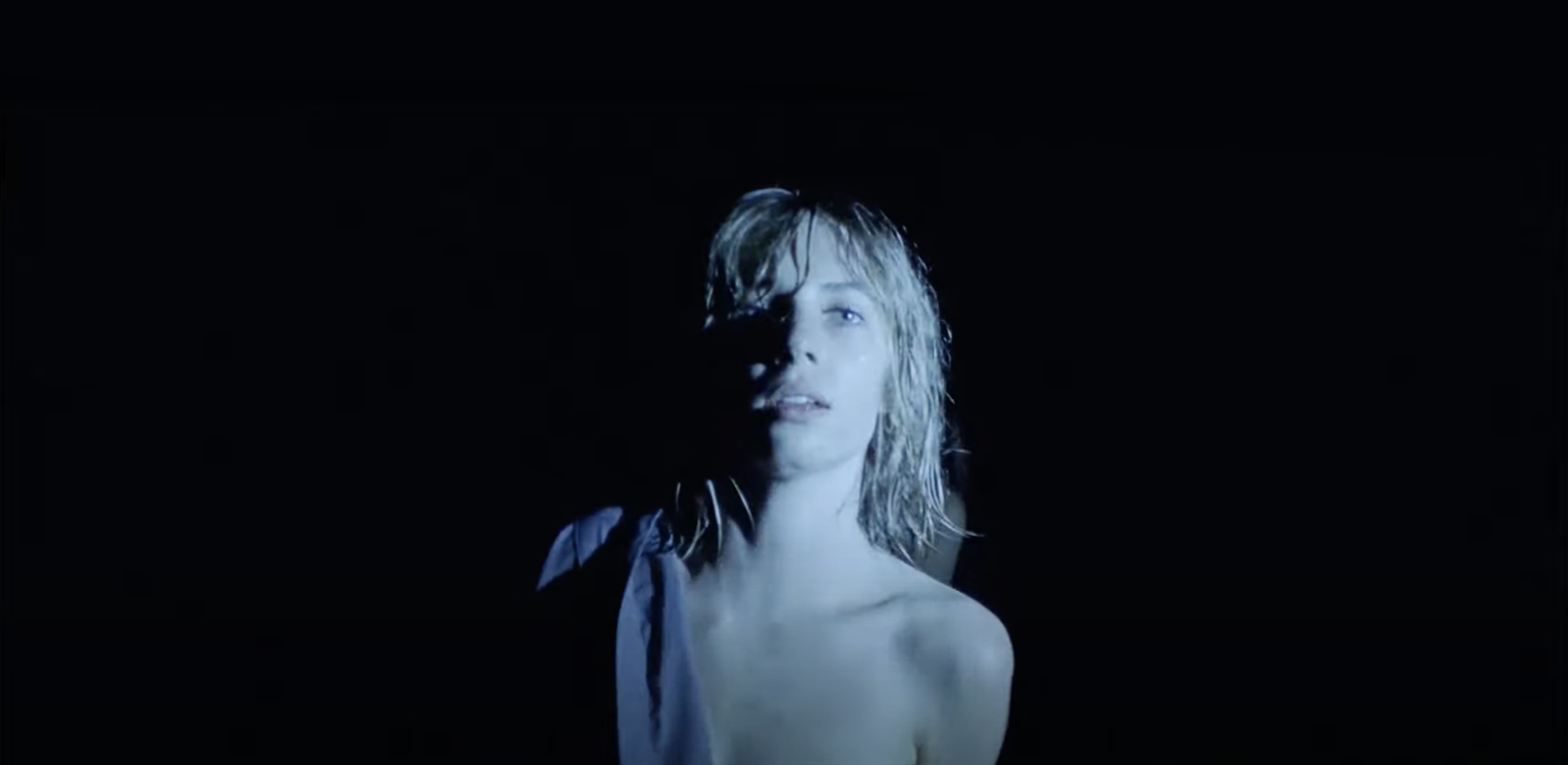 Maya Hawke standing topless in the dark with a shirt draped over her shoulder.